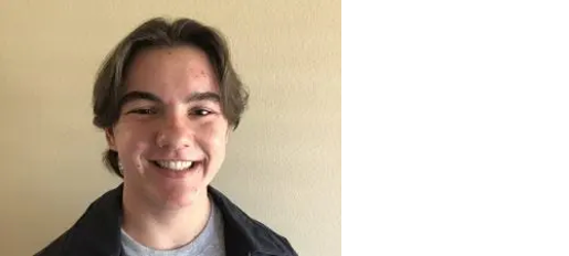 A young person with short brown hair and a light complexion smiles at the camera, standing in front of a plain beige wall.
